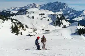 Deux skieurs équipés de leurs skis échangent au sommet d'une piste enneigée, avec une vue imprenable sur les montagnes alpines